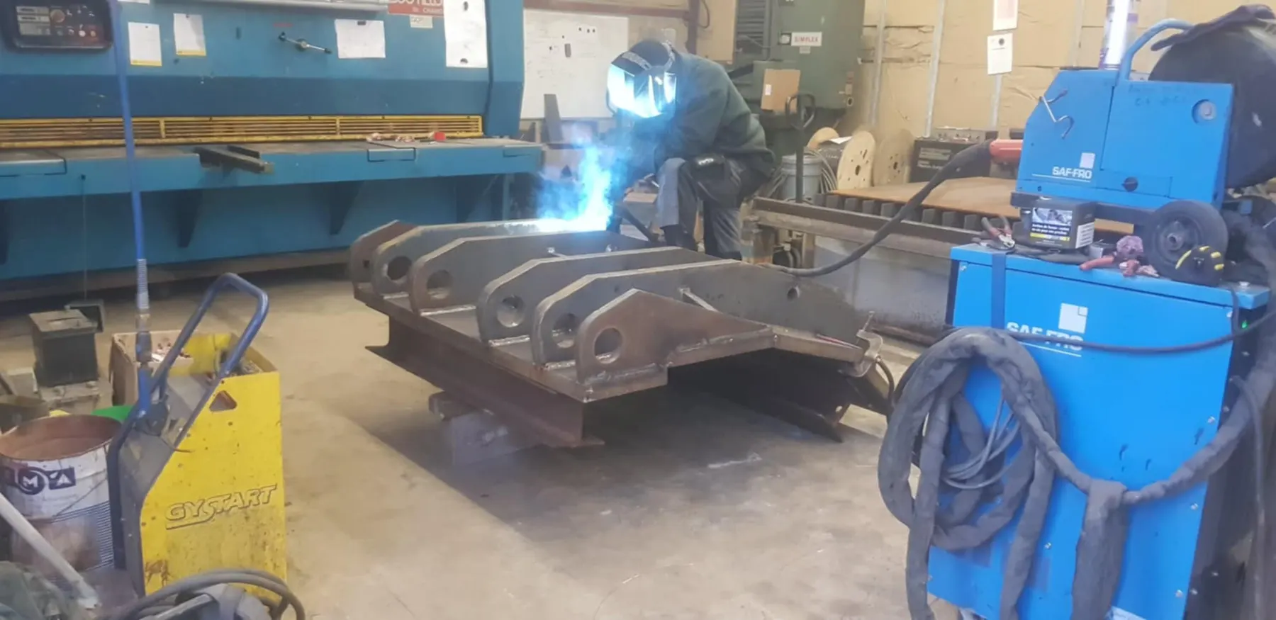 Soudure du châssis à l'atelier, soudeur casqué penché sur la pièce, poste à souder au premier plan